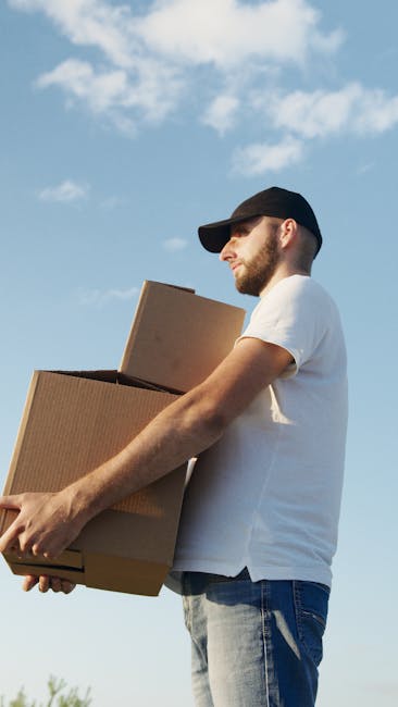 A man wearing a black cap, white t-shirt, and jeans is outdoors under a partly cloudy sky during daylight, carrying two cardboard boxes, with one box held close to his chest and the other balanced on top of it. He appears to be involved in a home relocation or furniture transport process, indicative of packing and moving activities. Behind him, a light-colored van with an open rear cargo area is visible, with the vehicle parked on a paved driveway or street, suggesting the loading or unloading phase of a house removal service. Inside the van, several cardboard boxes and wrapping materials, such as plastic stretch film, are arranged, indicating completion of packing tasks. The scene captures a professional move, with the man preparing to load or unload moving boxes to facilitate furniture transport and home relocation, while equipment like trolleys, straps, or blankets may be nearby to assist with safe handling, typical of the services offered by Man with Van Downham.