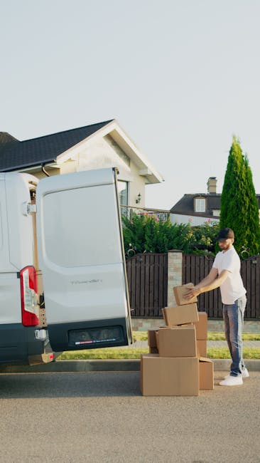 A man in casual clothing stands on a residential street next to a large, white moving van with its side door open. He is placing cardboard boxes onto the pavement, which are arranged in a sequence ready for loading or unloading. The interior of the van shows part of its empty cargo space, with a glimpse of packing materials possibly made of cardboard and paper. The background features a suburban house with a sloped roof, a garden with tall green trees, and a clear sky, indicating daylight. The scene appears to depict the home relocation or packing process, with the man assisting in furniture transport and moving logistics, supported by professional removals services such as those offered by Man with Van Downham.