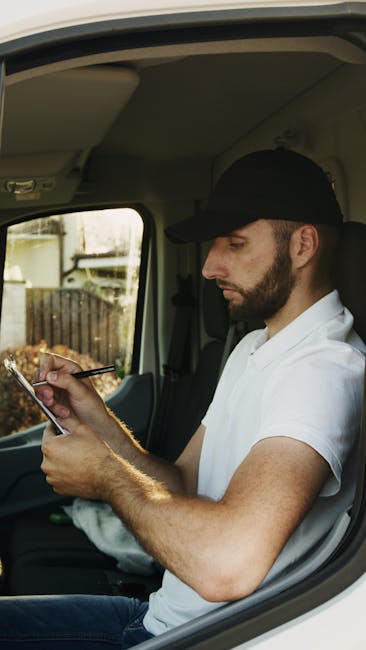 A man wearing a black cap, white t-shirt, and jeans is outdoors under a partly cloudy sky during daylight, carrying two cardboard boxes, with one box held close to his chest and the other balanced on top of it. He appears to be involved in a home relocation or furniture transport process, indicative of packing and moving activities. Behind him, a light-colored van with an open rear cargo area is visible, with the vehicle parked on a paved driveway or street, suggesting the loading or unloading phase of a house removal service. Inside the van, several cardboard boxes and wrapping materials, such as plastic stretch film, are arranged, indicating completion of packing tasks. The scene captures a professional move, with the man preparing to load or unload moving boxes to facilitate furniture transport and home relocation, while equipment like trolleys, straps, or blankets may be nearby to assist with safe handling, typical of the services offered by Man with Van Downham.