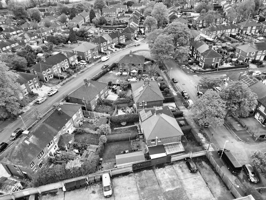 An aerial view in black and white showing a residential neighbourhood with closely spaced houses, some with front gardens and driveways, and vehicles parked along the streets. The image captures the rooftops, trees, and pathways, with a focus on a specific property that appears to be in the process of moving. Outside this property, there are several cardboard boxes, wrapped furniture items, and packing materials indicating packing and home relocation activity. A person from Man with Van Downham can be seen loading or unloading items into a van parked at the curb, with the van partially visible at the bottom of the image. The scene depicts an efficient loading process on the pavement, supported by additional moving equipment such as trolleys and straps that might be visible nearby. Surrounding houses and streets suggest a typical suburban environment, with the context reflecting the logistics involved in furniture transport, packing, and moving services within residential areas, consistent with Bromley Council skip and waste rules for Downham moves.