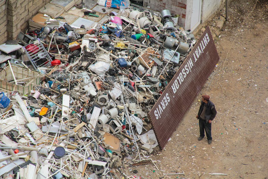 A man wearing a dark jacket and brown hat stands on a dirt surface next to a large pile of assorted household items, including plastic containers, metal pipes, electronic appliances, and miscellaneous