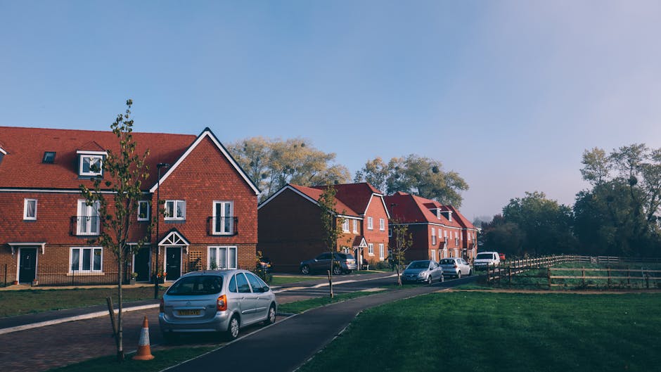 A row of modern red-brick residential houses located on a suburban street in the Downham Estate (BR1) area, with the front facades featuring pitched roofs, white-framed windows, and small front gardens. Several vehicles, including a grey hatchback, a black SUV, and a white van, are parked along the curbside on the paved road. In the foreground, a silver station wagon is parked next to a small, leafless tree with a traffic cone placed nearby, indicating a possible loading zone or moving activity. The scene is set during daytime with clear weather, soft shadows cast on the greenery and pavement, and a backdrop of trees with autumn foliage. The image contextualizes a typical residential environment where professional removals by Man with Van Downham occur during home relocations, with the street setup optimized for loading and unloading furniture and packed boxes, some wrapped in plastic or cardboard, in preparation for a house move.