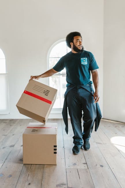 A man with dark curly hair and a beard is inside a bright, spacious room with white walls and wooden flooring, holding a cardboard box with red packing tape in his right hand. He is wearing a navy blue t-shirt with a light blue logo on the chest and dark trousers, standing beside two additional cardboard boxes, one on the floor and one being moved onto it, indicating a home relocation or furniture transport process. The room features a large arched window allowing natural light to illuminate the space, with no furniture or other objects visible, suggesting an early stage of packing or moving preparations. The overall scene reflects a professional removal or moving service environment, with the man representing a member of [COMPANY_NAME] involved in packing, loading, or organizing items for a house move, aligned with the context of the Downham Estate (BR1) moving guide on [PAGE_TITLE].