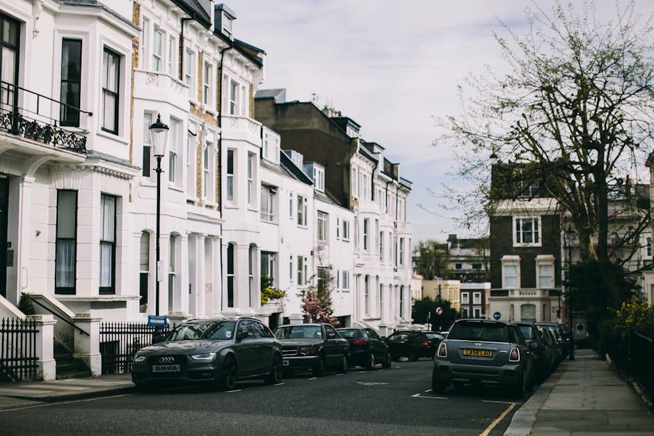 A row of modern red-brick residential houses located on a suburban street in the Downham Estate (BR1) area, with the front facades featuring pitched roofs, white-framed windows, and small front gardens. Several vehicles, including a grey hatchback, a black SUV, and a white van, are parked along the curbside on the paved road. In the foreground, a silver station wagon is parked next to a small, leafless tree with a traffic cone placed nearby, indicating a possible loading zone or moving activity. The scene is set during daytime with clear weather, soft shadows cast on the greenery and pavement, and a backdrop of trees with autumn foliage. The image contextualizes a typical residential environment where professional removals by Man with Van Downham occur during home relocations, with the street setup optimized for loading and unloading furniture and packed boxes, some wrapped in plastic or cardboard, in preparation for a house move.