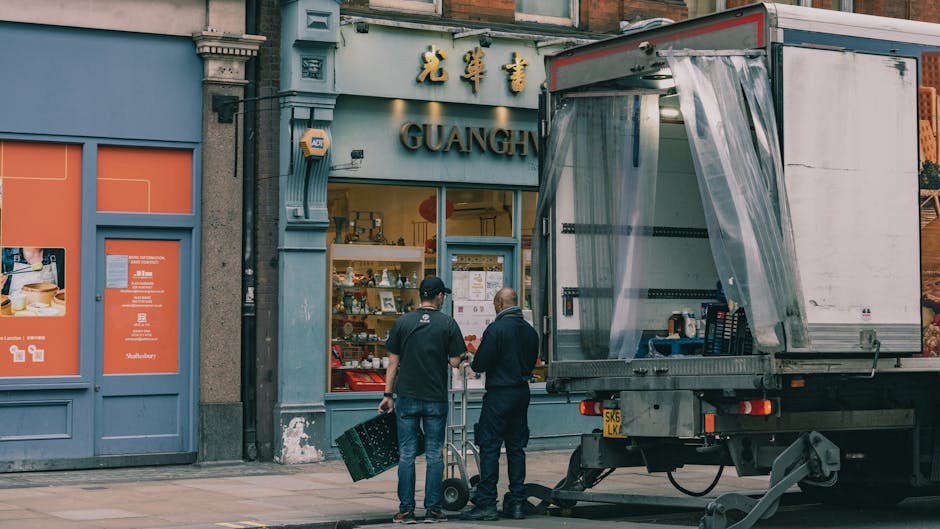 Two men are engaged in a home relocation or furniture transport activity outside a shop on Downham High Street, as part of professional removals services by Man with Van Downham. One man, dressed in a black shirt and jeans, is holding a black plastic crate, while the other, in a dark jacket and pants, is standing beside a hand truck with a green base and black handle. They are positioned near a loading bay area with a partially opened large delivery truck, which features a transparent plastic curtain on its rear door. The truck is parked on the sidewalk with its rear elevated, ready for loading or unloading supplies. Inside the shop window, various items are visible, indicating retail or specialty store offerings. The scene is set in daylight, with natural light illuminating the street environment, and the surrounding buildings exhibit a traditional storefront facade typical of UK high streets. This visual captures the logistical process involved in furniture or packing and moving activities central to house removals, supported by the professional services of Man with Van Downham.