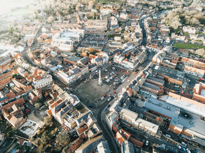 An aerial view of Downham High Street showing a busy town square with a tall central monument surrounded by a paved area. Nearby are parking lots filled with cars, closely lined streets with a mix of shops, residential buildings, and commercial properties. The scene includes vehicles moving along the roads, with some parked on the street and in parking areas. The buildings have pitched roofs, mostly with brick or tile exteriors, and the area is bordered by greenery and additional residential neighborhoods in the distance. The overall environment illustrates a typical UK town centre, captured in daylight, highlighting the urban layout and commercial activity relevant to house removals and transportation services, as managed by Man with Van Downham.