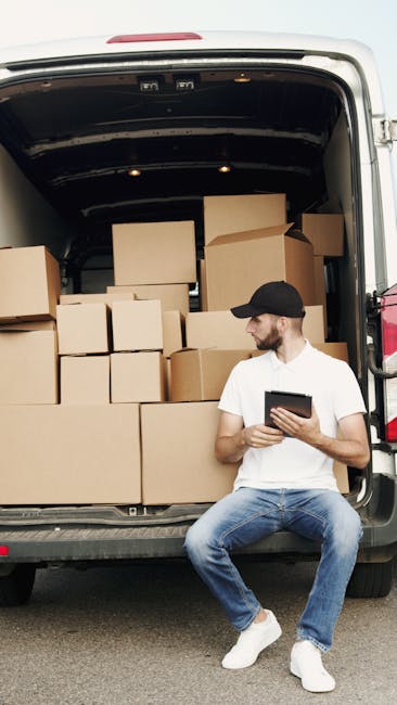 Two men are engaged in a home relocation process involving furniture transport outside a residential property. One man stands on the paved driveway, wearing casual clothing and white sneakers, looking