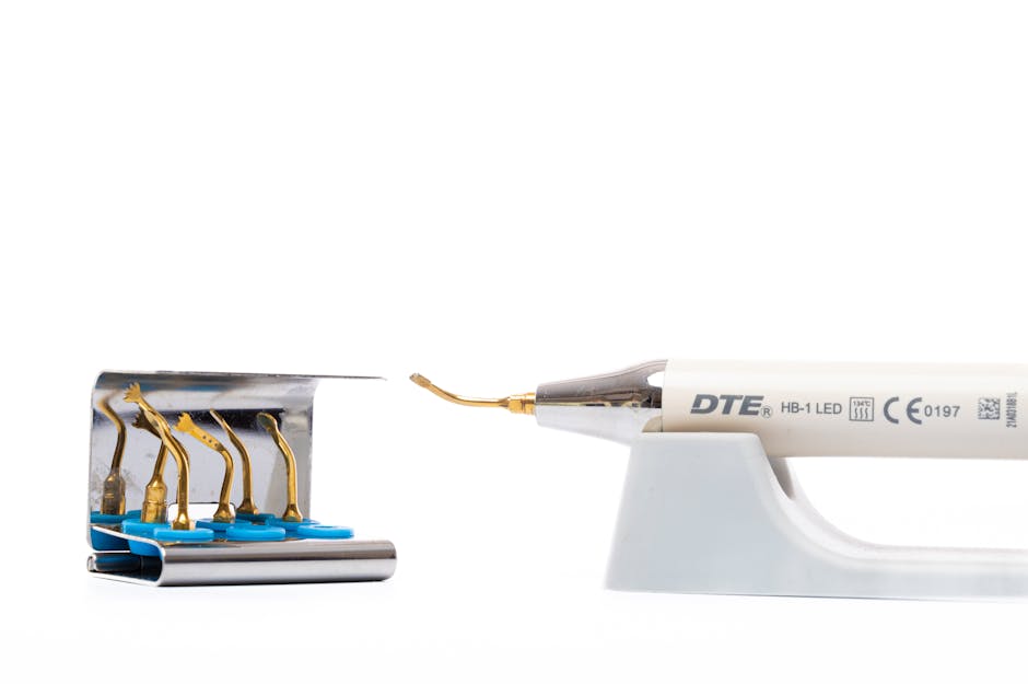 A close-up view of a soldering iron with a metal tip, positioned on a white plastic stand, connected to a power source, with the cord extending out of the frame. To the left, there is a small metal bl