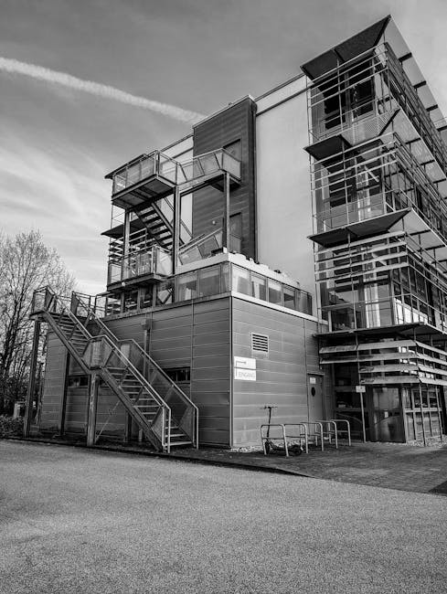 A black and white photograph of a modern multi-story building with external metal staircases and numerous balconies, situated at the corner of the structure. The building features large glass windows and metal framing, with sections of the facade composed of horizontal panels. Snow is visible on some parts of the balconies, indicating a winter setting. The sky is clear with visible contrails, and there are leafless trees in the background. In the foreground, a paved area with a bicycle rack and a sign on the building reading 'ENTRANCE' can be seen. The image captures the building's exterior design, likely a commercial or office premises, with clear details of the stairs, balconies, and structure. Man with Van Downham would ensure careful planning for any home relocation or furniture transport to or from such a property, considering its external access features.