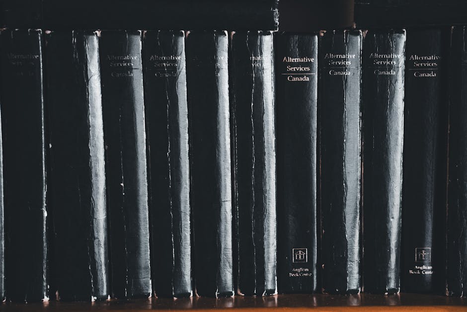 A row of black hardcover books with white and orange text on the spines, neatly aligned on a wooden shelf. The books appear to be organized in numerical or thematic order, with titles such as 'Alternative Services Canada' and other similar labels visible. The spines show some signs of wear, and the shelf is indoors, illuminated by soft lighting. While not directly related to house removals or packing, this image may suggest the importance of organized space or documentation during a home relocation process. The setting reflects a clean, structured environment typical of a professional moving or packing service like Man with Van Downham, which specialises in furniture transport and loading processes tailored for house moves. The image’s neutral tone emphasizes orderliness and careful handling, key aspects of efficient removals.