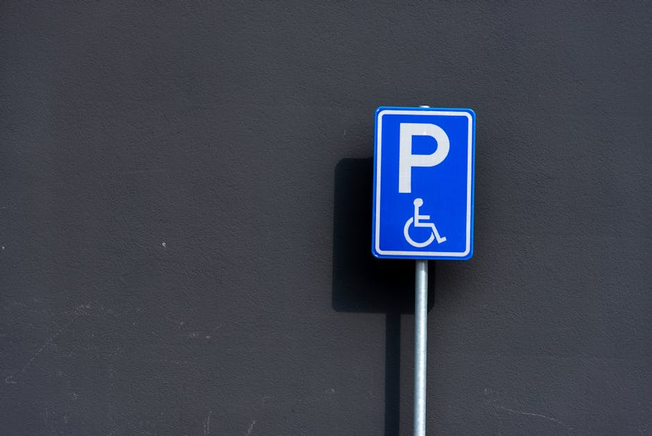 A blue and white disability parking sign mounted on a metal pole, positioned against a dark grey exterior wall. The sign features a large white letter 'P' and a wheelchair symbol below, indicating accessible parking. The shadow of the pole falls on the wall, illuminated by natural outdoor lighting. The scene depicts an outdoor area suitable for home relocation services, with the sign marking a designated parking space for disabled individuals, relevant for planning furniture transport and loading during a move. Man with Van Downham occasionally references such signage as part of moving logistics and parking arrangements for efficient house removals, ensuring accessible parking compliance during the loading process.