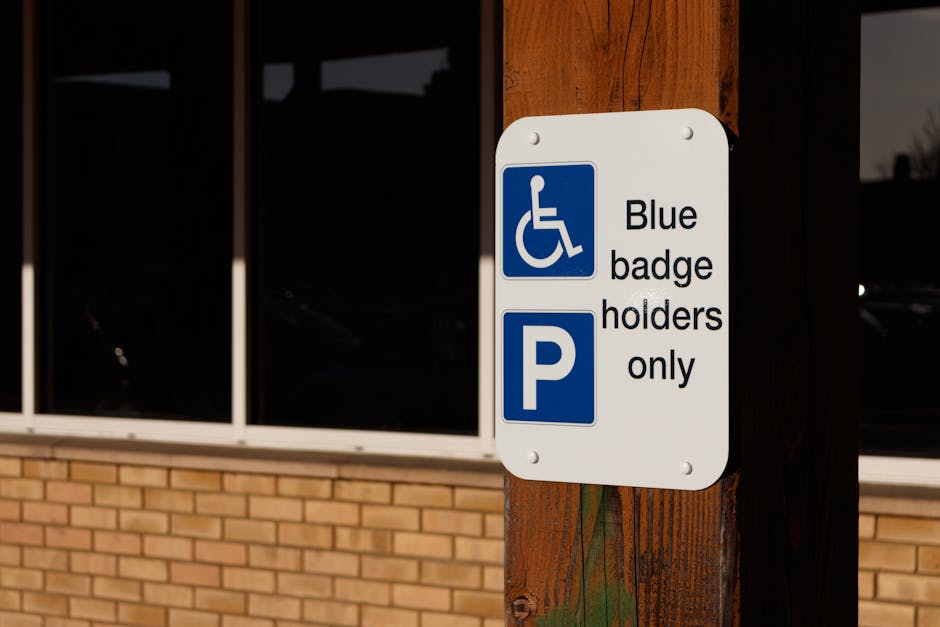 A white rectangular parking sign mounted on a dark brown wooden post outdoors in front of a brick building with large glass windows. The sign features a blue wheelchair symbol at the top, indicating accessibility, and the words 'Blue badge holders only' written in black text next to a blue square with a white 'P', signifying parking restrictions. The environment appears well-lit, suggesting daytime, with reflections visible in the windows. The sign demonstrates facilities relevant for disabled parking, which may be useful for house removals or moving services planning in Lewisham. Man with Van Downham uses such signage awareness as part of their logistics in home relocation and furniture transport, ensuring compliance with parking regulations during loading and unloading operations near residential or commercial properties.