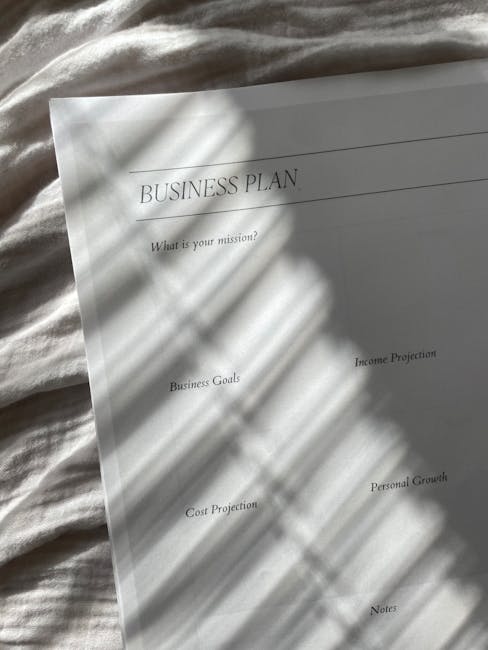 A close-up of a printed business plan document lying on a light-colored fabric surface, with natural sunlight casting shadows from nearby window blinds across the page. The document includes headings