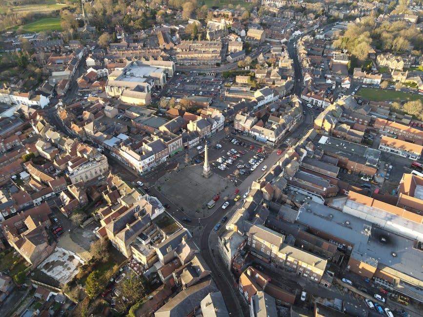 An aerial view of a small town showing a central town square with a tall monument, surrounded by tightly packed terraced houses and commercial buildings. In the town square, there are several parked cars and an open paved area with a few vehicles, possibly including a truck or van used for home relocation services. The surrounding streets are narrow and winding, with some parked cars along the sides. Adjacent to the square, residential properties with small gardens or paved yards are visible, and the overall environment appears calm with abundant greenery at the edges of the town. The image highlights the dense urban layout that Man with Van Downham might operate within during furniture transport and packing and moving processes, especially in busy areas where local logistics can impact move timings.