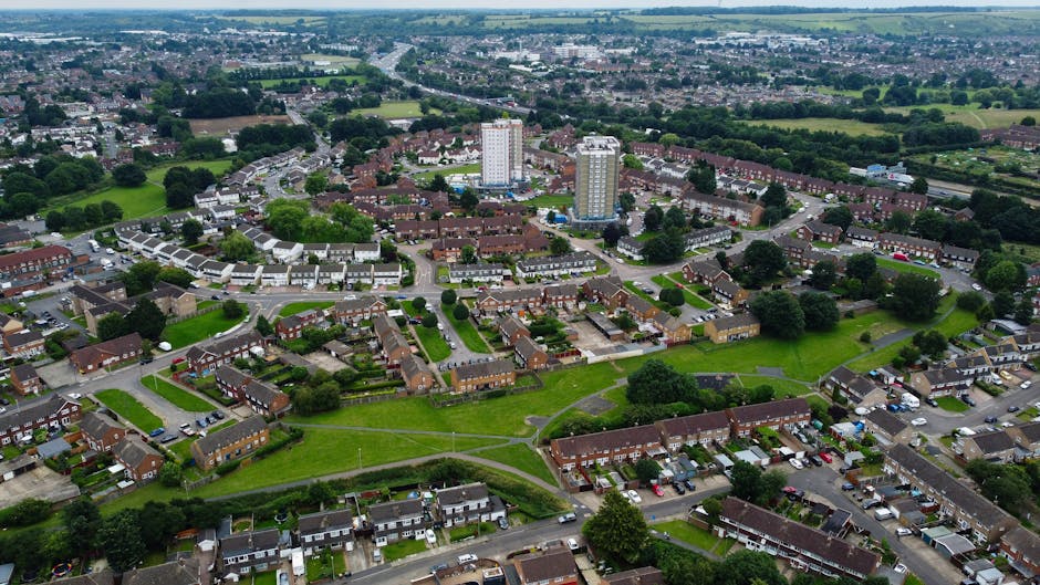 An aerial view of a residential neighbourhood in Downham showing a mix of terraced houses, semi-detached homes, and low-rise apartment buildings. The image captures a central green park area with walking paths and surrounding mature trees. Several streets are lined with parked cars, and the area appears quiet and well-kept. In the foreground, a row of houses with front gardens can be seen, with some vehicles parked along the curbs. The background reveals more scattered houses and open green spaces extending towards the horizon, under a cloudy sky. This urban landscape illustrates the type of area where house removals and furniture transport services might take place, with clear access to residential roads suitable for moving trucks. Man with Van Downham can assist with home relocation and moving logistics in such environments, ensuring efficient packing and loading processes for a smooth move in Downham.